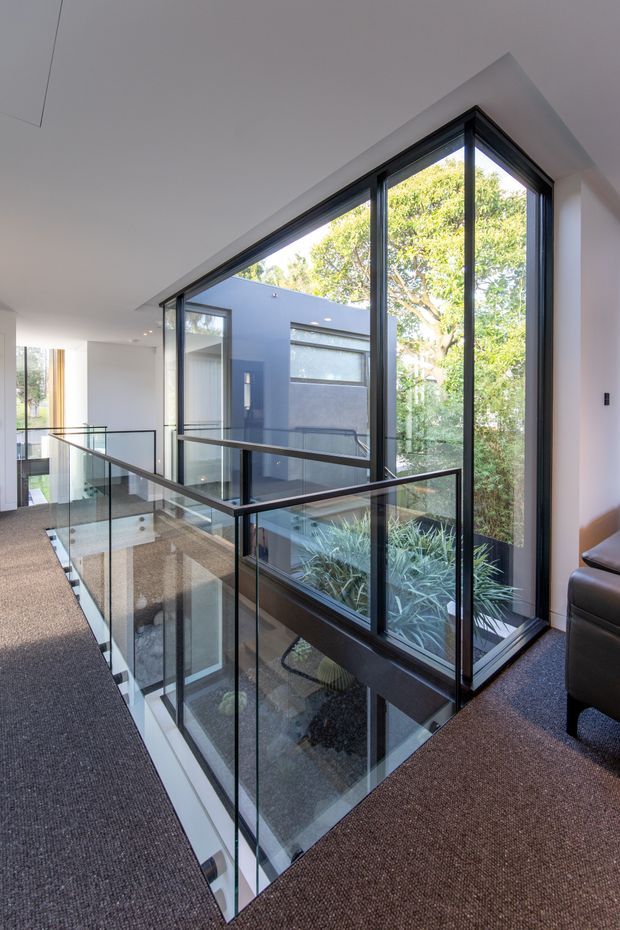 Interior view of a contemporary upper-level landing or hallway overlooking a lower floor through a glass balustrade. The space features full-height black-framed glazing, a dramatic angled roofline, white walls and ceiling, dark carpeted floors, and views into a landscaped internal courtyard or garden. A partial leather sofa is visible at the right edge.