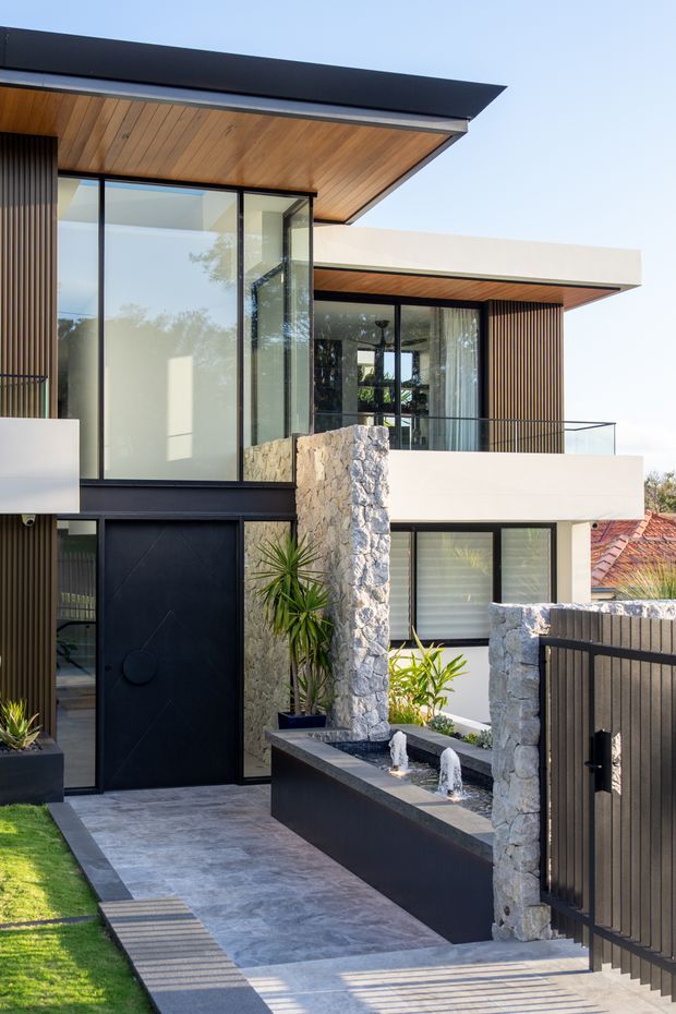 A contemporary multi-level residential facade viewed from the front entry. The house features crisp white rendered volumes, dark metal-framed glazing and doors, vertical dark screening, a projecting flat roof with a warm timber-lined soffit, and rough natural-stone walls. A paved entry path leads past a narrow black water feature with small fountains, integrated planting, and a dark metal pedestrian gate.