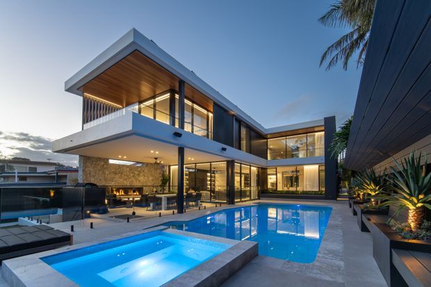 Twilight exterior view of a large contemporary multi-storey residence arranged around a landscaped pool courtyard. The home features crisp cantilevered volumes, extensive floor-to-ceiling glazing, dark vertical and horizontal cladding, a projecting upper balcony, warm timber soffits, and an illuminated swimming pool with a raised spa in the foreground. Palm planting and built-in garden beds frame the property.
