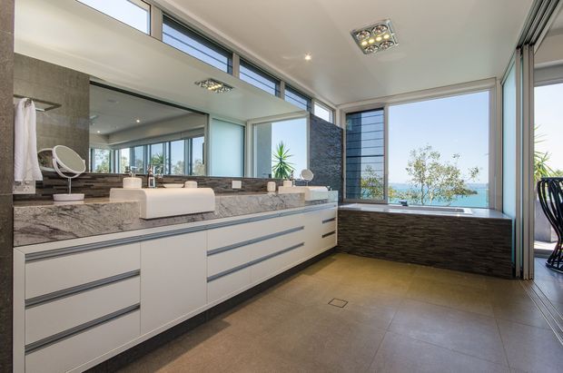 Contemporary, spacious bathroom with a long double vanity featuring white integrated basins, sleek handleless cabinetry, and a mottled stone benchtop. Large mirrored surfaces, expansive windows, clerestory glazing, dark textured wall cladding, and large-format neutral floor tiles create a bright, refined interior. A freestanding bathtub is positioned beside the windows, with a glimpse of ocean or blue water beyond.