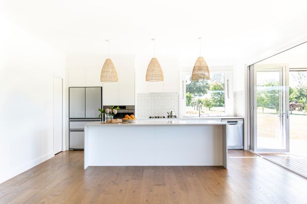 Bright contemporary kitchen with a large white island, integrated dark cabinetry and appliances, a pale patterned tile splashback, timber flooring, woven pendant lights, and full-height glazed doors opening to the outdoors.