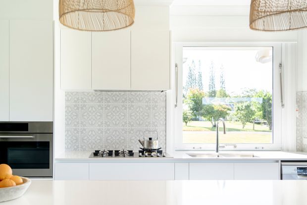 Bright contemporary kitchen with white cabinetry, a patterned grey-and-white tiled splashback, a gas cooktop, integrated oven, stainless-steel sink and tap beneath a large window. Woven pendant lights hang above a glossy white island or benchtop, with a leafy suburban outlook beyond.