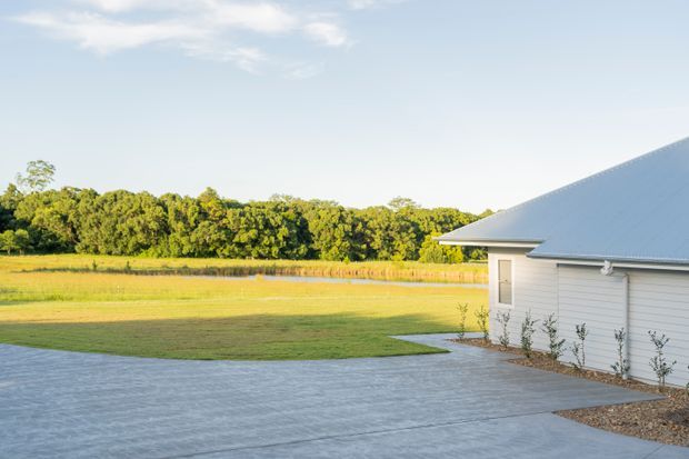 Exterior view of a single-storey building with pale horizontal weatherboard cladding and a broad light metal pitched roof. A large concrete-paved forecourt borders an expansive grassy rural or semi-rural landscape with mature trees.