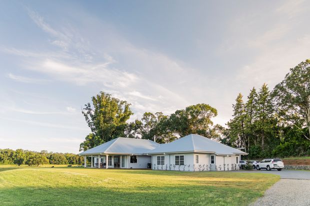 A wide exterior view of a low, single-storey white house set in a spacious landscaped rural property. The home has a light-coloured hipped roof, broad eaves, covered verandah-like outdoor areas, white cladding, and extensive surrounding lawn and mature trees. A driveway and parked vehicles are visible at the side.