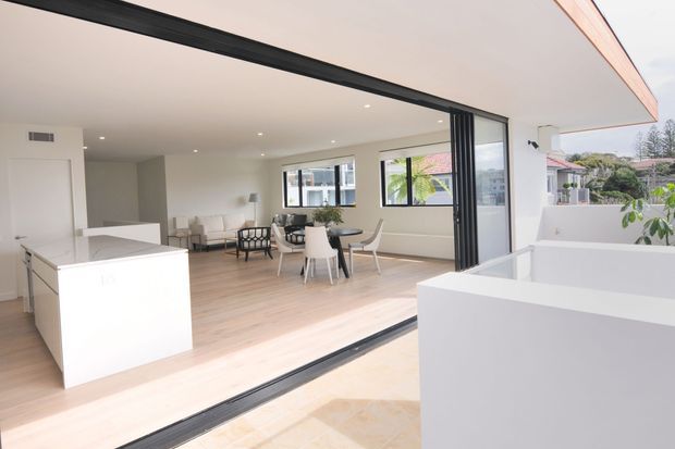 Bright contemporary open-plan kitchen and dining area opening onto an outdoor terrace through large black-framed sliding glass doors. The space features a white island, pale timber flooring, white walls and cabinetry, recessed ceiling lighting, and a neutral dining setting. White terrace planters and neighbouring residential buildings are visible outside.