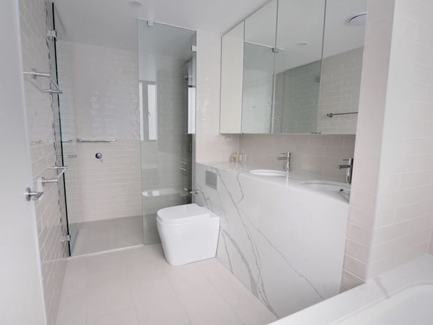 Bright contemporary bathroom featuring a frameless glass shower, freestanding-style white toilet, white bathtub, mirrored wall cabinetry, pale tiled walls and floor, and a white marble-look vanity with grey veining. Chrome fixtures and fittings provide subtle contrast.