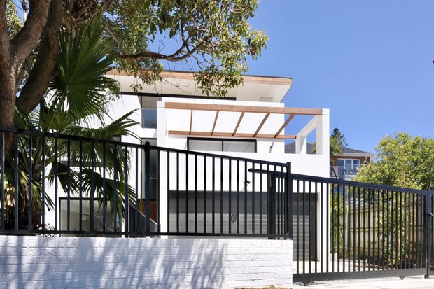 Street-facing view of a contemporary white multi-level house with a flat roof, broad glazing, a projecting pergola or awning structure, and a dark vertical-bar security fence above a low white masonry wall. Mature trees and palms frame the property.
