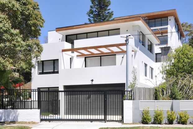 A contemporary multi-storey residential home viewed from the street, with a crisp white rendered facade, extensive dark-framed glazing, warm timber roof and pergola elements, balconies, and a black metal driveway gate. The property is set among mature trees and includes a landscaped front boundary.