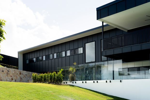 Exterior view of a contemporary multi-level residence with dark vertical cladding, broad low-pitched roof planes, projecting upper-level balcony, extensive glazing, glass balustrades, and a stone retaining wall set within a landscaped sloping lawn.