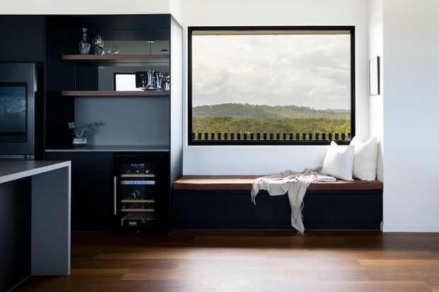 Contemporary kitchen and dining nook with dark built-in cabinetry, integrated wine storage, a black-framed picture window, and a built-in window seat overlooking a green landscape. Warm timber flooring contrasts with the crisp white walls and dark joinery.