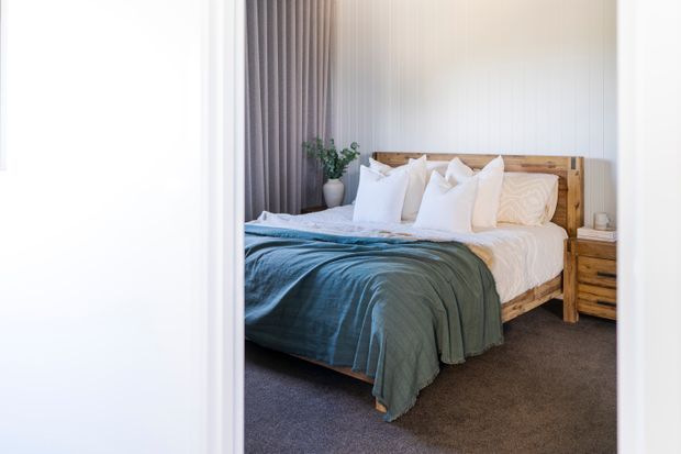 Bright, calm bedroom viewed through a doorway, featuring a timber bed with a natural live-edge style headboard, white layered bedding, a muted blue-green throw, grey carpet, full-height grey curtains, and vertical white wall panelling. A timber bedside table and potted plant add warmth and texture.