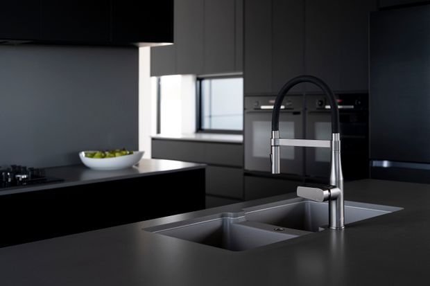A sleek, dark contemporary kitchen focused on a large stainless-steel undermount double sink and tall gooseneck mixer tap set into a dark benchtop. Black and charcoal cabinetry surrounds integrated appliances, with a kitchen island and a bright horizontal window providing contrast.