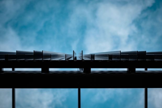 Abstract architectural detail viewed from below, featuring a dark horizontal structure with repeating angled metal or glass panels silhouetted against a vivid blue, clouded sky. The image emphasizes geometric lines, rhythm, contrast, and structural form; no interior room or clearly identifiable building facade is visible.