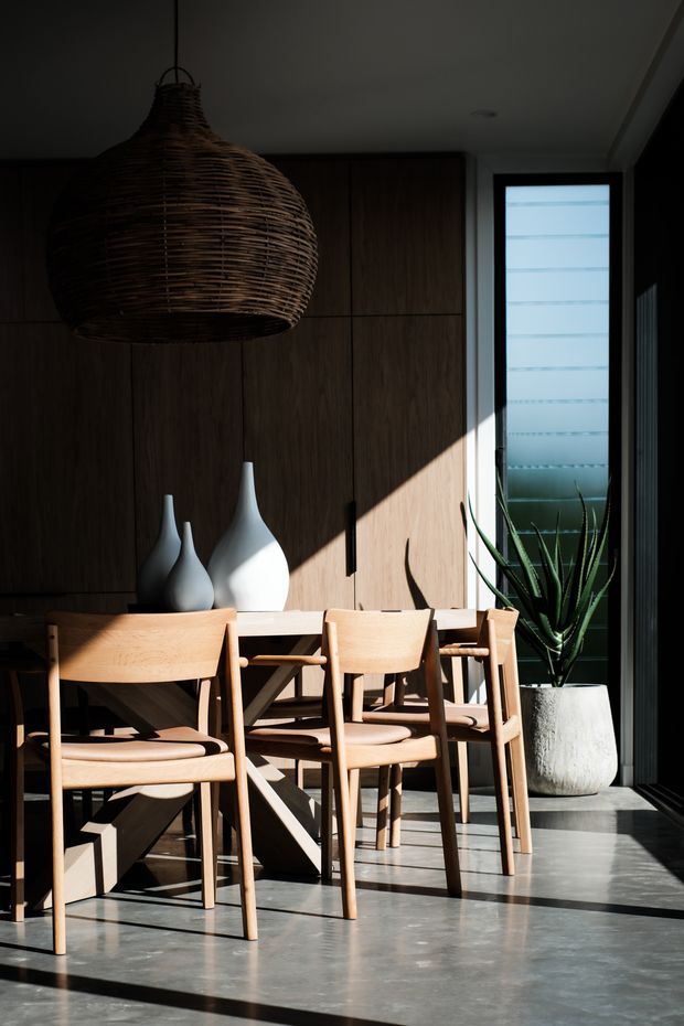 A sunlit dining area with a warm timber dining table and matching wooden chairs, set against dark wood wall cabinetry. A large woven pendant light hangs overhead, with sculptural white ceramic vases on the sideboard and a tall potted plant beside a narrow frosted window. Strong diagonal sunlight creates dramatic shadows across the timber furniture and concrete-look floor.
