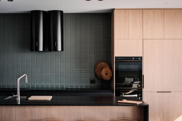 Contemporary kitchen with a black stone benchtop, dark green-grey tiled splashback, pale timber cabinetry, integrated black double oven, and two sculptural black cylindrical rangehoods. A slim gooseneck tap and minimal accessories contribute to a refined, modern composition.