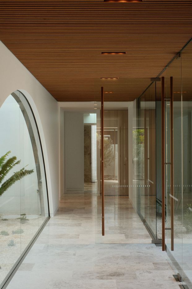 A refined residential hallway or entry passage with a polished pale stone floor, white walls, extensive frameless glass partitions, and slim vertical timber door handles. The ceiling is lined with closely spaced warm timber battens and recessed linear lighting. A large arched glazed opening on the left brings in natural light and reveals planted landscaping, while glass doors and screens on the right create a light-filled, gallery-like circulation space.