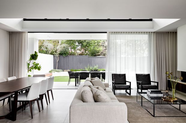 Bright contemporary open-plan living and dining area with a large neutral sectional sofa, black-framed coffee table, black lounge chairs, and a dark timber dining table with white upholstered chairs. Full-height sheer curtains and glazing open to a landscaped courtyard, while a broad overhead opening brings in additional natural light.