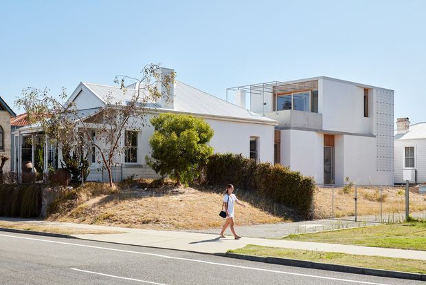 Street view of a residential property combining an older single-storey gabled cottage with a crisp contemporary two-storey addition. The original home has pale weatherboard cladding, a pitched metal roof, chimney and traditional proportions, while the extension features white rendered walls, a flat roof, upper-level glazing, a balcony/terrace and a perforated screen wall. The front garden is dry and sparsely landscaped, with a footpath and suburban street in the foreground.