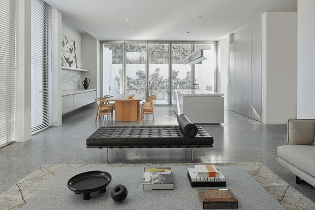 A bright, open-plan contemporary interior combining living, dining, and kitchen areas. The room features polished grey flooring, white walls and cabinetry, full-height glazed windows with white blinds, a streamlined kitchen island, a timber dining table, and a black tufted daybed in the foreground. The restrained neutral palette is complemented by sculptural furniture and minimal artwork.