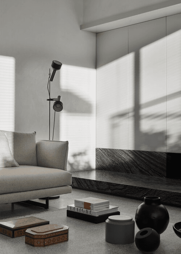 A refined contemporary living room with a low cream upholstered sofa, sculptural black floor lamp, dark veined stone media console and wall treatment, and a curated arrangement of black ceramic vessels and stacked books on a low coffee table. Strong sunlight casts geometric window shadows across the pale walls and joinery, creating a calm, high-contrast atmosphere.