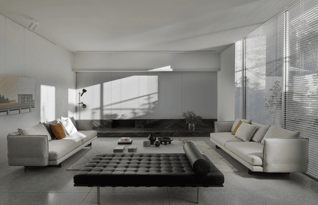 A refined modern living room with a neutral grey and white palette. Two light upholstered sofas face a low black tufted ottoman and central coffee table, with a dark stone or marble feature wall and fireplace/media recess behind them. Full-height windows with horizontal blinds provide strong filtered daylight, while minimal decor and clean architectural lines create an open, sophisticated interior.