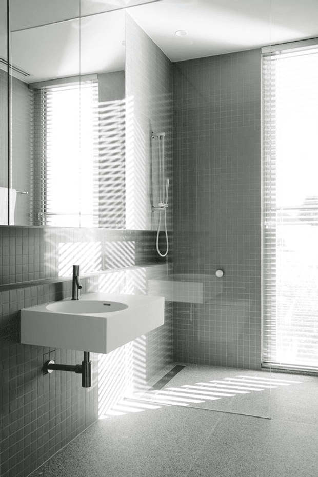 A bright, minimalist contemporary bathroom with a wall-mounted white rectangular basin, exposed chrome plumbing, large mirrored wall panels, and a walk-in shower with a slim handheld shower rail. Small-format grey square tiles cover the walls, while the floor is a light grey textured finish. Strong sunlight filters through venetian blinds, creating striped shadows across the tiled surfaces and floor.