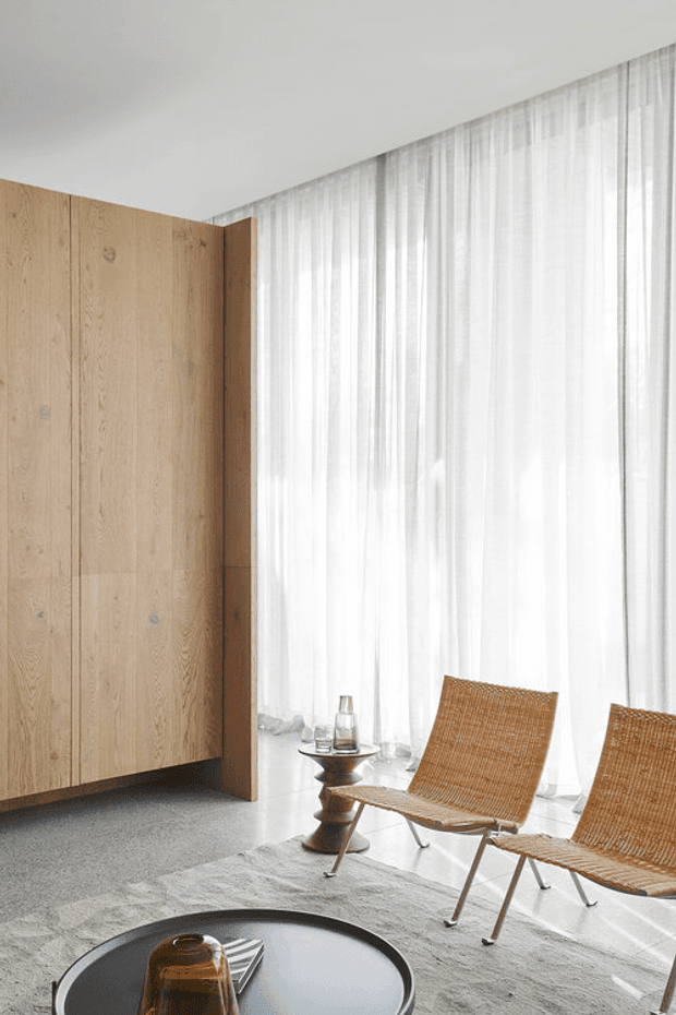 A bright minimalist lounge or living room with a floor-to-ceiling sheer-curtained window, pale timber built-in cabinetry or wall panelling, two woven lounge chairs, a low round dark coffee table, and a neutral textured rug. The space has a calm, restrained palette and abundant diffused natural light.