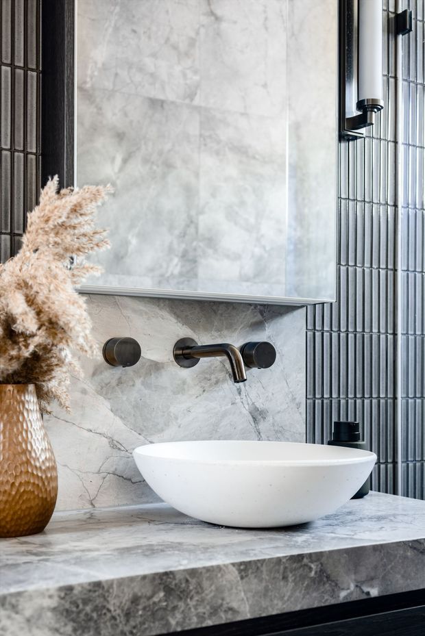 A refined bathroom vanity vignette featuring a white vessel basin on a grey veined stone benchtop, wall-mounted dark metal tapware, a large frameless mirror, and vertically ribbed dark wall tiles. A textured woven vase with dried pampas grass adds a warm decorative accent.