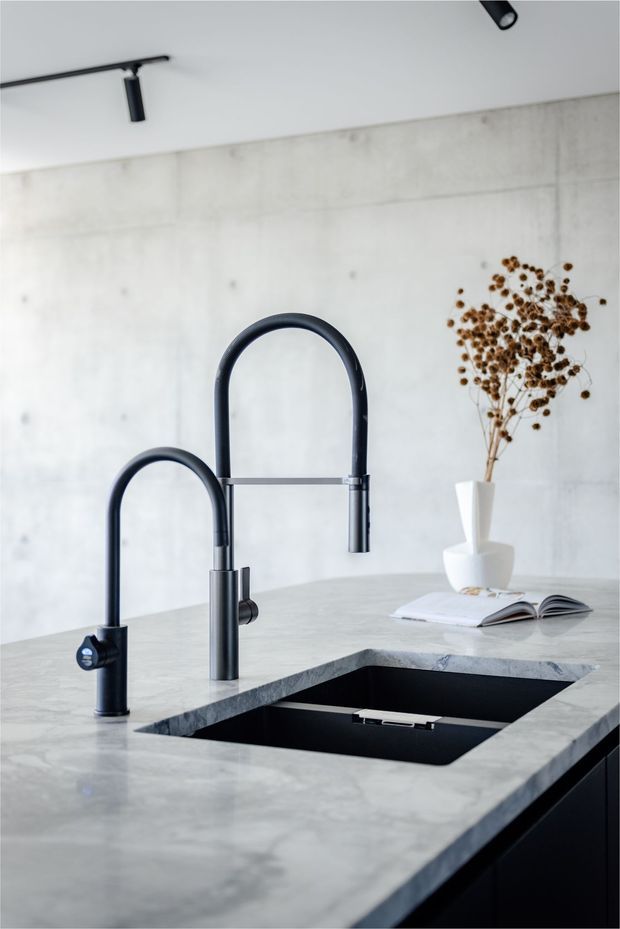 Close-up view of a contemporary kitchen island featuring a pale grey stone benchtop, integrated black undermount sink, and two tall matte-black gooseneck tap fixtures. The island has dark cabinetry below, with a light concrete-look wall and black track lighting in the background. Minimal styling includes a white vase with dried branches and an open book.