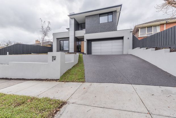 Street-facing exterior view of a contemporary two-storey home with a flat, projecting roofline, dark grey brick upper facade, white rendered walls, large windows, and an integrated double garage. A broad asphalt driveway rises from the footpath, bordered by low rendered walls and dark fencing.