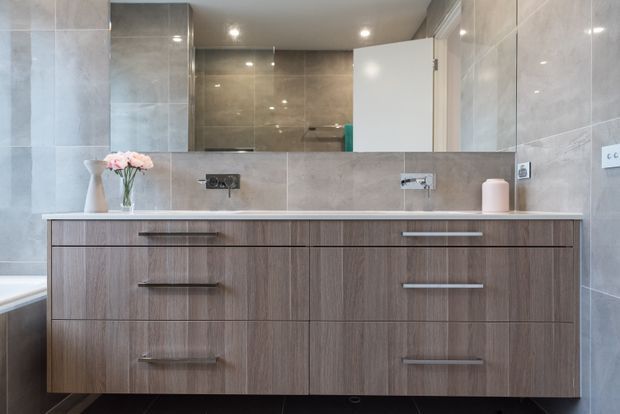 Contemporary bathroom vanity with a wide timber-look floating cabinet, long horizontal metal handles, white countertop, and twin wall-mounted basins and tap fittings. Large grey porcelain wall tiles continue around the room, with a mirrored wall cabinet or splashback above the vanity, recessed ceiling lights, and a partially visible bathtub at left. Soft pink flowers and a small accessory add a subtle decorative touch.