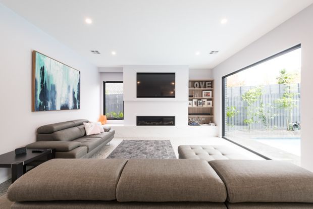 Bright contemporary living room with a large modular sectional sofa and ottoman seating, a wall-mounted television above a built-in fireplace, open shelving, a large abstract artwork, and expansive glazed doors opening to an outdoor area. The room has white walls and ceiling, dark-framed glazing, recessed lighting, and a neutral grey and taupe palette.