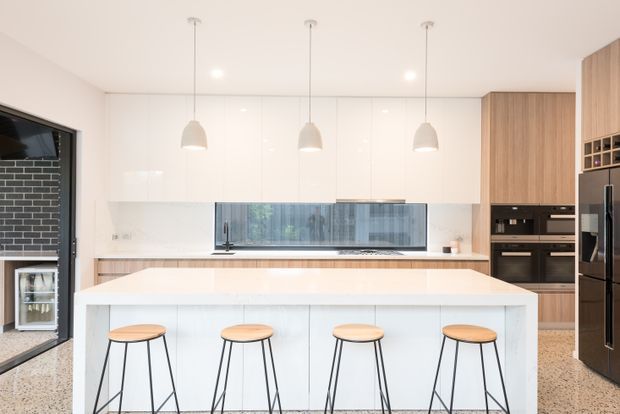 Bright contemporary kitchen with a large white island and four slim black-framed stools, glossy white full-height cabinetry, warm timber cabinetry, integrated appliances, a reflective splashback, and three pendant lights. A fireplace with dark brickwork is visible at the left edge, and terrazzo-style flooring extends through the space.