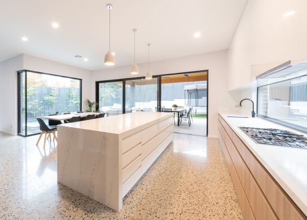 Bright, spacious contemporary kitchen with a large white stone island, timber cabinetry, integrated gas cooktop, terrazzo-style flooring, pendant lights, and extensive black-framed glazing opening toward an outdoor dining area.