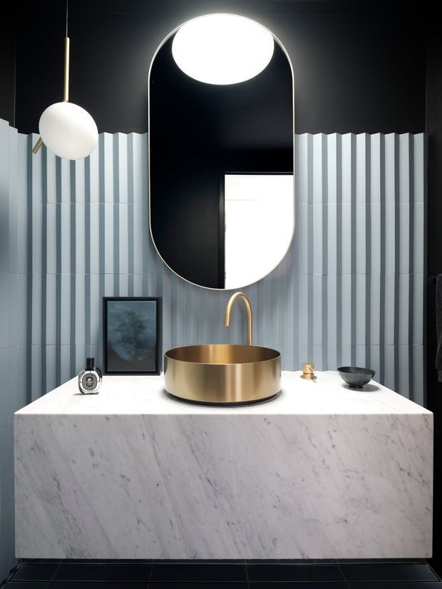 A dramatic contemporary bathroom vanity featuring a freestanding round brass basin and matching curved faucet on a substantial white marble-look vanity. The wall behind is clad in vertically fluted pale blue-grey tiles, contrasted with a dark ceiling and a large rounded-rectangle black-framed mirror. Sculptural pendant lighting and restrained black-and-brass accessories create a refined, high-contrast scheme.