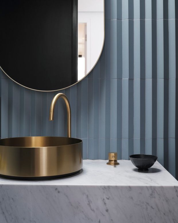A dramatic contemporary bathroom vanity featuring a round brushed-gold vessel basin, curved matching faucet, and small gold tap set on a thick white marble benchtop. The wall is clad in vertically arranged blue-grey glazed tiles with alternating projecting ribs. A large round black-framed mirror with a fine metallic edge dominates the upper left, while a dark decorative bowl sits on the counter.