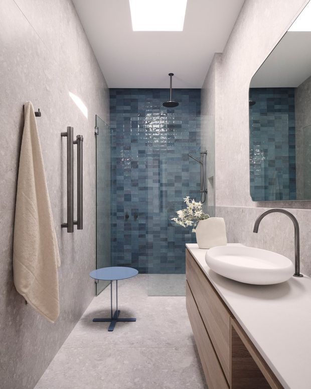 A narrow, contemporary bathroom with a walk-in shower at the rear, lined in varied blue rectangular tiles. The space has pale textured walls and flooring, a large wall mirror, a timber vanity with a white countertop, a white vessel basin, and dark metal fixtures. A skylight provides bright natural illumination, complemented by a black ceiling shower and heated towel rail.