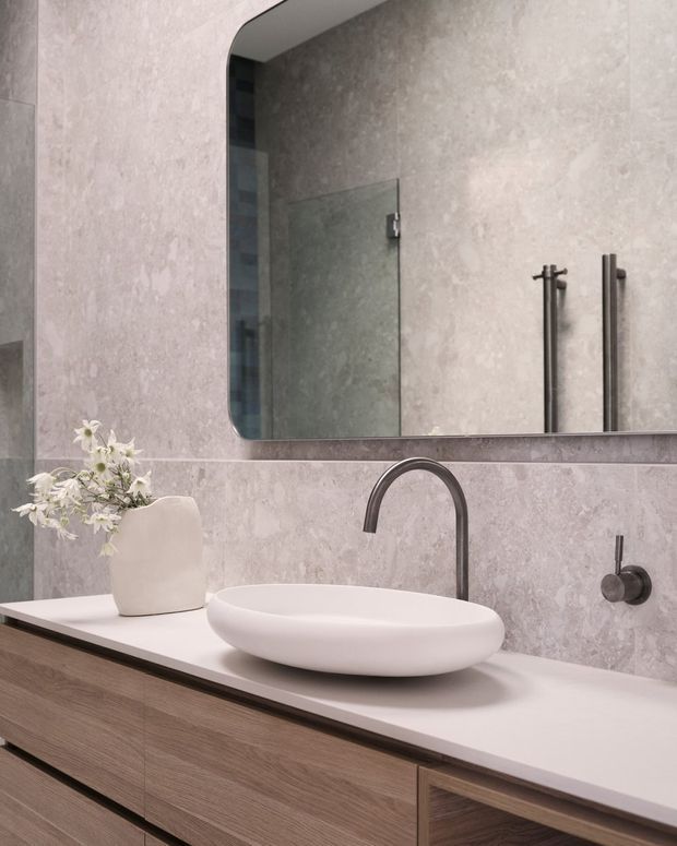 Contemporary bathroom vanity with a white vessel basin on a pale stone benchtop, warm timber-grain cabinetry, grey textured wall tiling, dark brushed-metal tapware, and a large rounded-corner mirror. A glass shower enclosure and heated towel rail are visible in the reflection and background.