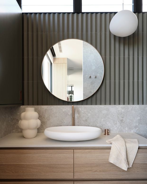 A refined contemporary bathroom vanity area featuring a floating timber-look drawer cabinet, pale stone benchtop and splashback, a white vessel basin, brushed brass tapware, a large round mirror, and dark vertically ribbed wall panelling. A sculptural pendant light and neutral accessories add a calm, tactile finish.