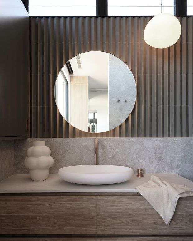A refined contemporary bathroom vanity with a floating timber-grain cabinet, pale stone countertop and backsplash, white vessel basin, round mirror, and sculptural pendant light. The wall behind the mirror features dark vertical ribbed panelling, creating a strong textural contrast with the light stone and warm wood tones.