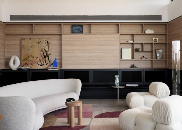 Contemporary living room with a large built-in timber wall unit incorporating open shelving, concealed storage, artwork niches and a long black media console. A curved cream sofa and sculptural rounded lounge chairs sit on a patterned rug, with small timber side tables and restrained decorative objects. The room has a warm, refined palette and integrated linear air-conditioning grille.