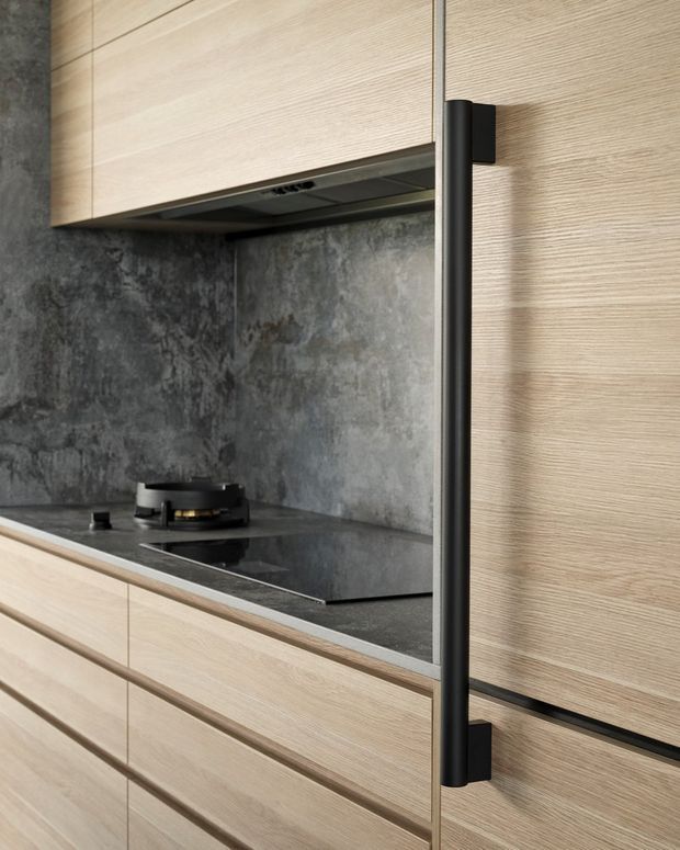 Close-up view of a contemporary kitchen with light wood-grain cabinetry, integrated black appliances and handles, a dark stone-look benchtop, and a mottled dark grey splashback. The image focuses on the cooktop, drawers, overhead cabinetry, and tall pantry-style door hardware.