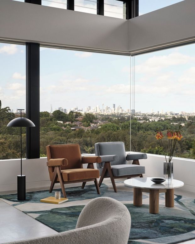A refined living room with expansive corner glazing overlooking a leafy suburban landscape and distant city skyline. The room features two mid-century inspired lounge chairs, a sculptural boucle chair in the foreground, a round coffee table, a dark abstract rug, and a minimalist black floor lamp. White walls, dark window frames, and high-level glazing create a bright, elevated interior with a strong indoor-outdoor connection.