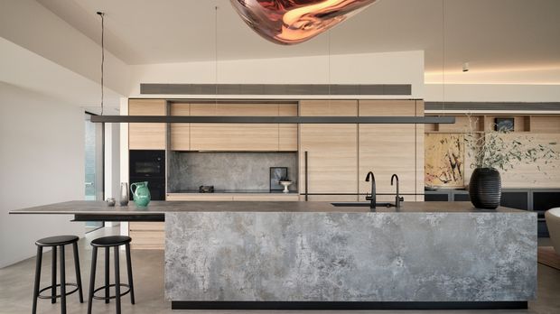 A refined contemporary kitchen dominated by a large island with a mottled grey stone or concrete-look front and dark benchtop. Full-height light timber cabinetry spans the rear wall, incorporating integrated appliances, open shelving and a dark sink and tapware. Black pendant lighting and stools provide contrast, while a sculptural reflective pendant is partially visible above. The space is open-plan and bright with a restrained neutral palette.