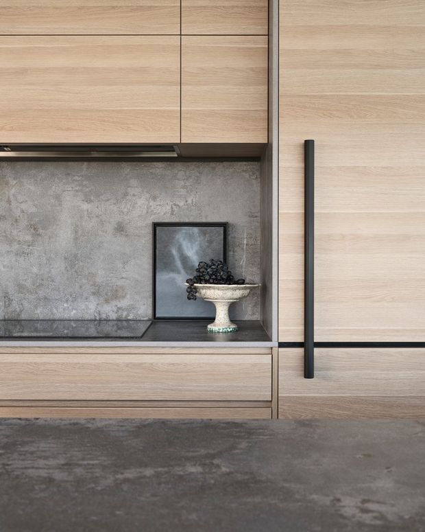 Close-up of refined built-in kitchen or storage joinery in pale wood tones, with flush horizontal cabinetry, a tall door with a long dark handle, a mottled grey concrete-like backsplash, dark benchtop, and a small decorative display niche. The composition is minimalist and detail-focused, highlighting the cabinetry and material contrasts.