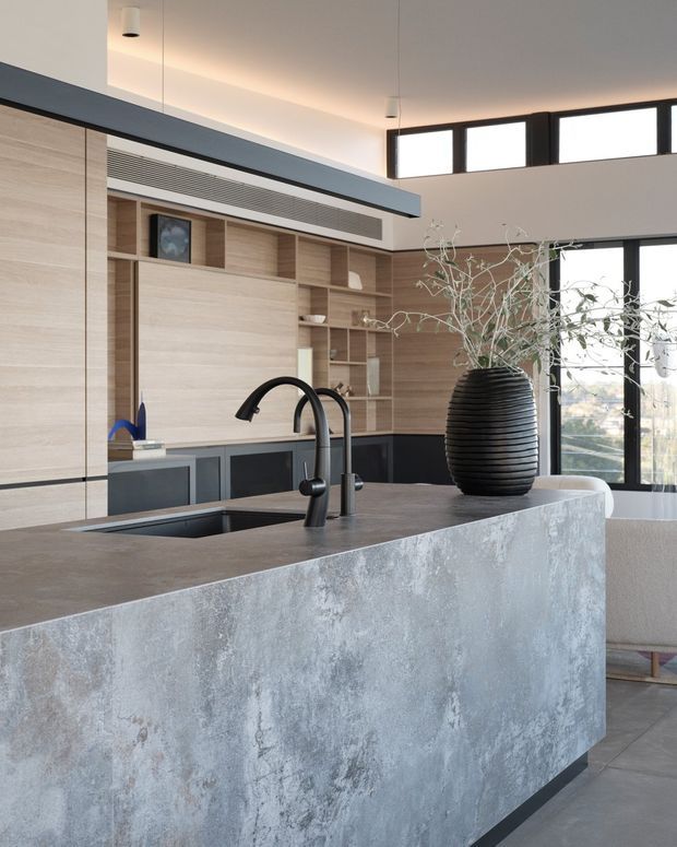 Contemporary open-plan kitchen dominated by a large grey textured island with an integrated sink and matte black tapware. The kitchen features light timber built-in cabinetry, dark lower cabinets, open shelving, integrated ventilation, and expansive black-framed clerestory and full-height glazing. A black ribbed vase sits on the island, while a partially visible upholstered seating area is beyond.