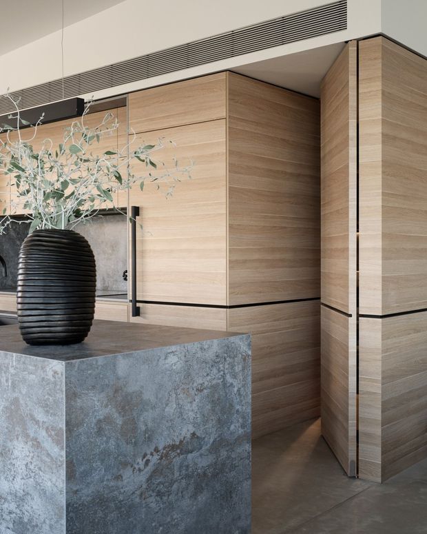 A refined contemporary interior focused on a large kitchen island and full-height timber-look cabinetry. The cabinetry has warm horizontal wood grain with dark shadow gaps and integrated detailing, while the island is clad in a blue-grey mottled stone surface. A sculptural black ribbed vase with pale branches sits on the island. A linear ceiling air-conditioning grille and restrained palette create a minimalist, high-end feel.