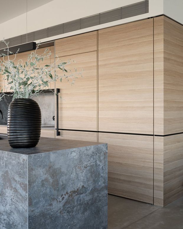 A refined contemporary kitchen interior featuring a large light timber wall of flush-panel cabinetry, a substantial grey stone island, dark integrated details, and a sculptural black ribbed vase with foliage. A linear ceiling air vent runs above the cabinetry, while concrete-toned flooring and a dark-framed opening reinforce the minimalist, material-focused design.
