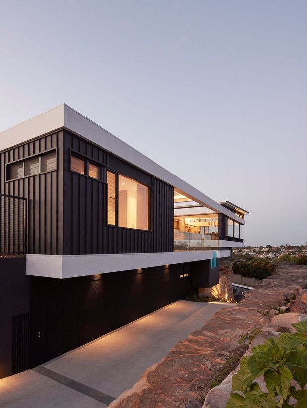 Dusk exterior view of a contemporary multi-level residence built into a sloping rocky site. The home features dark vertically ribbed cladding, crisp white rendered bands, extensive glazing, a projecting upper volume, recessed exterior lighting, and a driveway leading to a concealed garage. Warm interior light contrasts with the dark facade, while a residential suburb is visible in the distance.