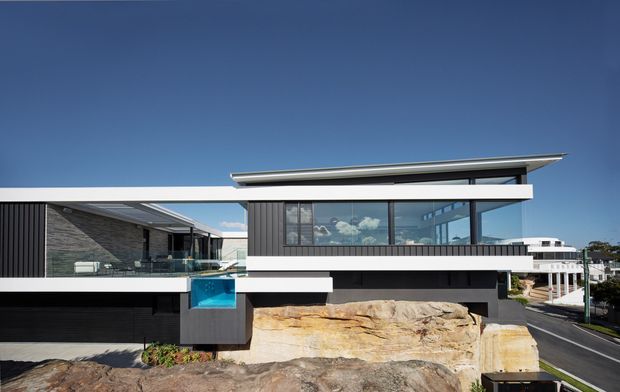 Exterior view of a contemporary multi-level residence built into a rocky sloping site. The house features strong horizontal white bands, dark vertical cladding, extensive glazing, cantilevered forms, and a prominent shallow roof plane. A small blue pool or water feature is visible at the front, with neighbouring buildings and a road in the background.